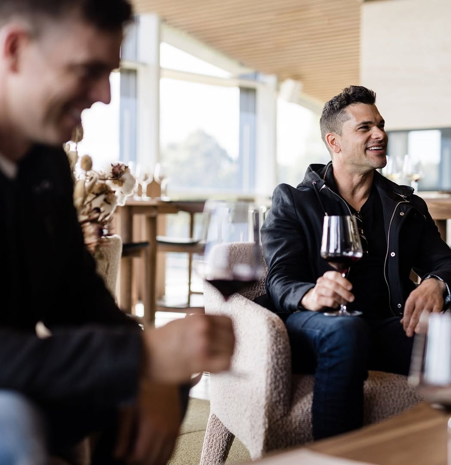 Two men sitting indoors, one holding a glass of red wine, with a blurred background.